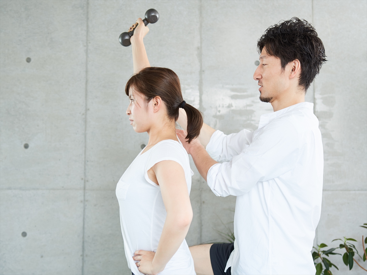 ダンベルを持つ女性の腕と肩の位置に手を添えて修正する男性トレーナー