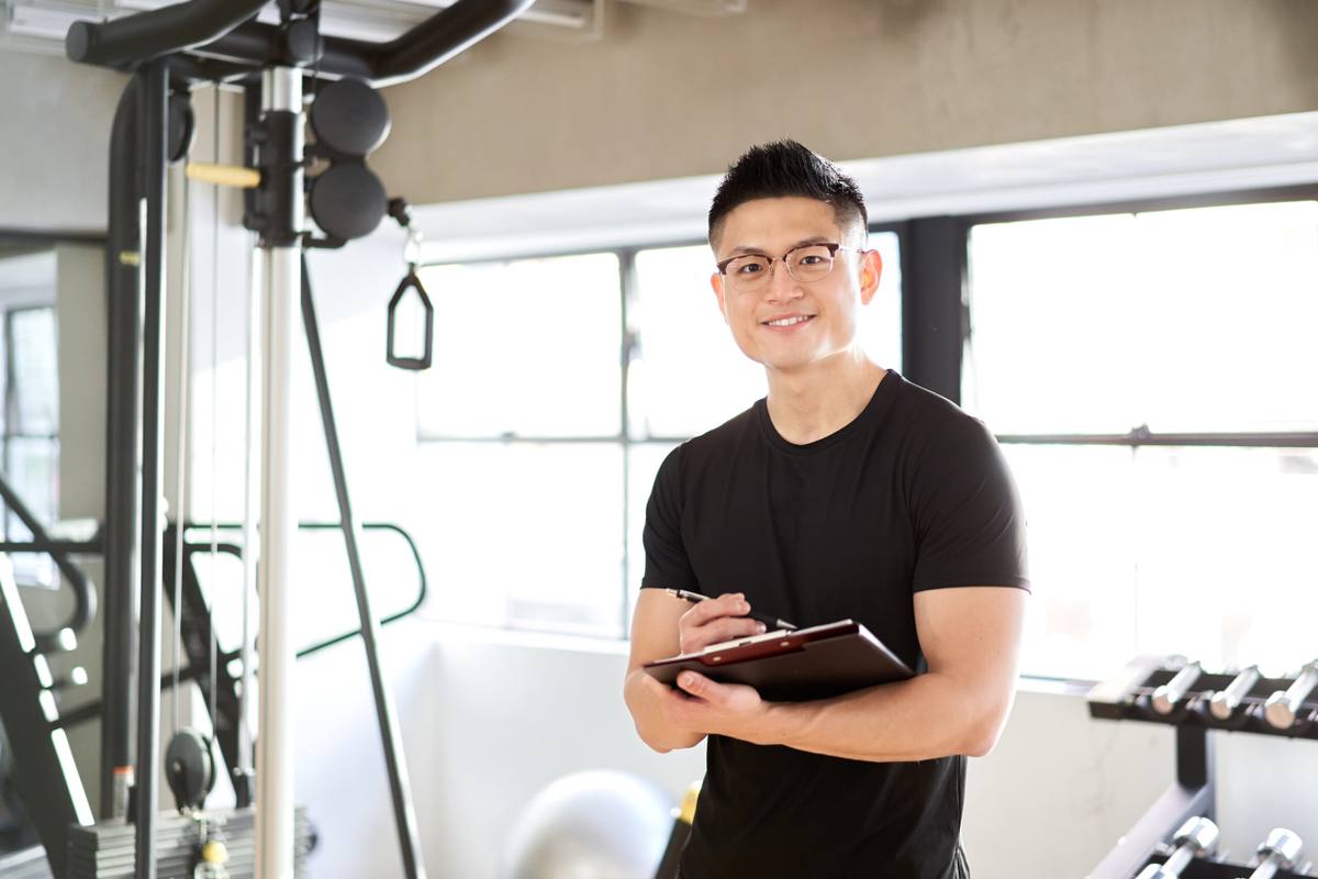 明るいジムで、黒いTシャツの男性トレーナーがクリップボードを持って立っている