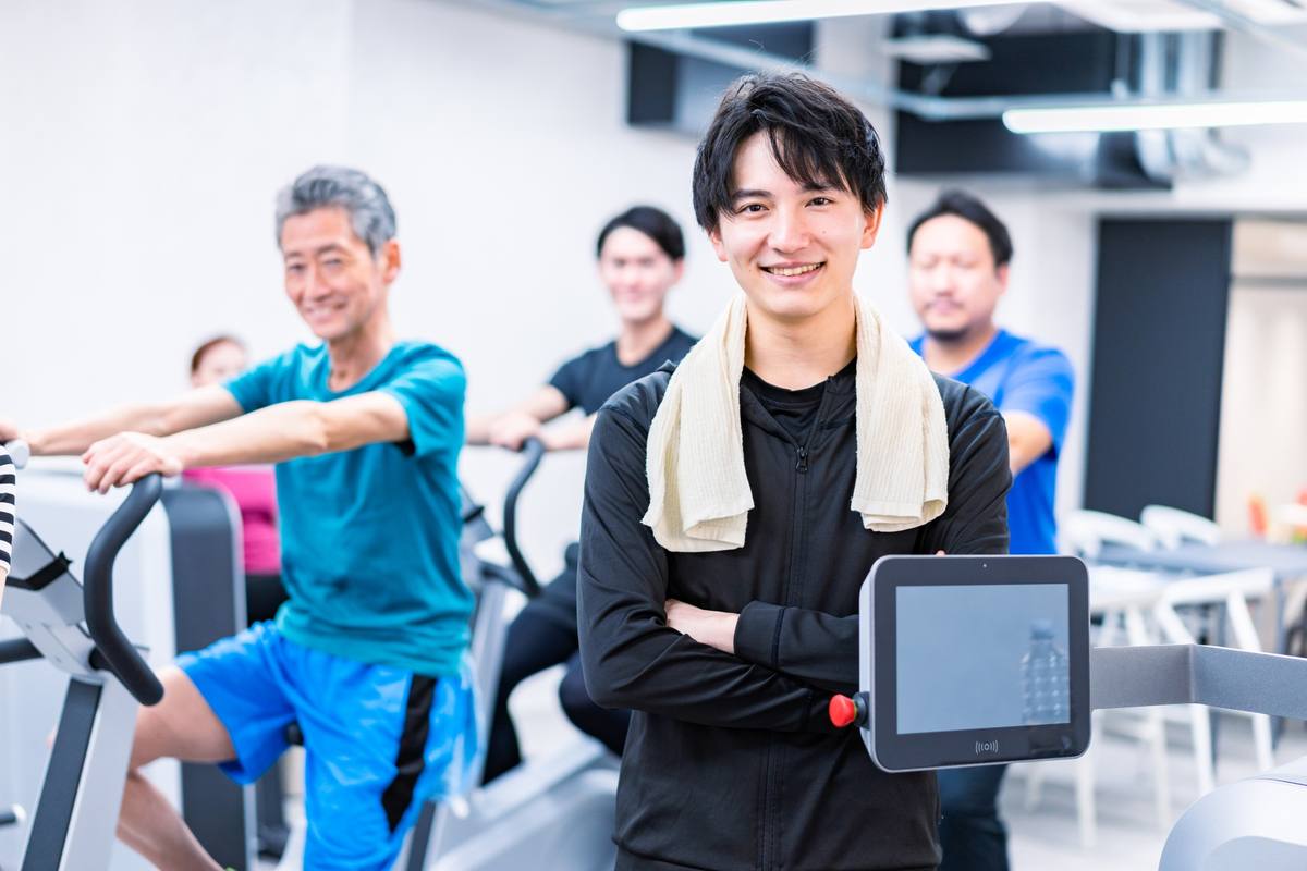 フィットネスクラブで運動する年配の男性を含む利用者と、タブレットを持つスタッフ