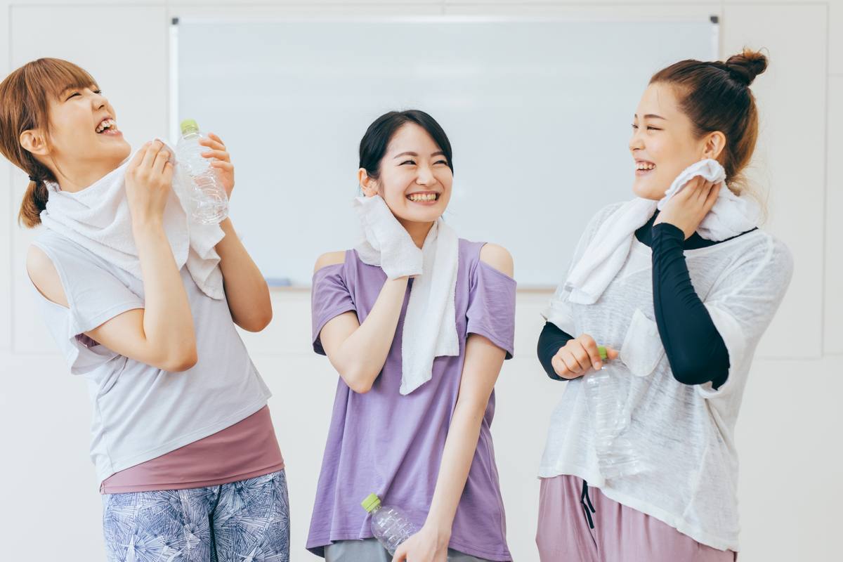 白い背景の前で、タオルを持った3人の女性が笑いながら話している様子