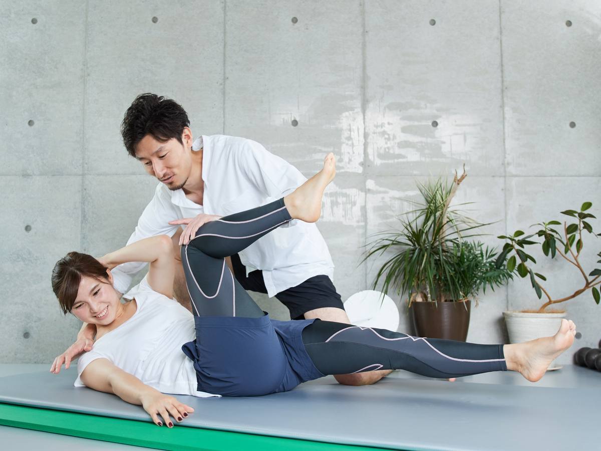 トレーナーが横向きに寝た女性の脚を持ち上げて伸ばす、二人組で行うストレッチの場面
