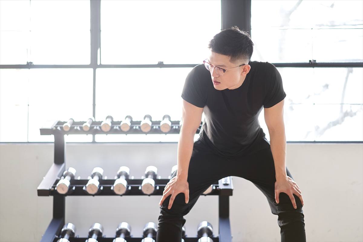 黒いトレーニングウェアの男性がダンベルラックの前で腰を落として構えているところ