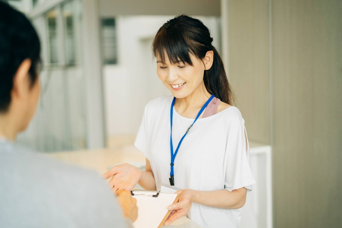 ジムの受付カウンターで、名札を付けた女性スタッフが来店者に笑顔で対応している様子