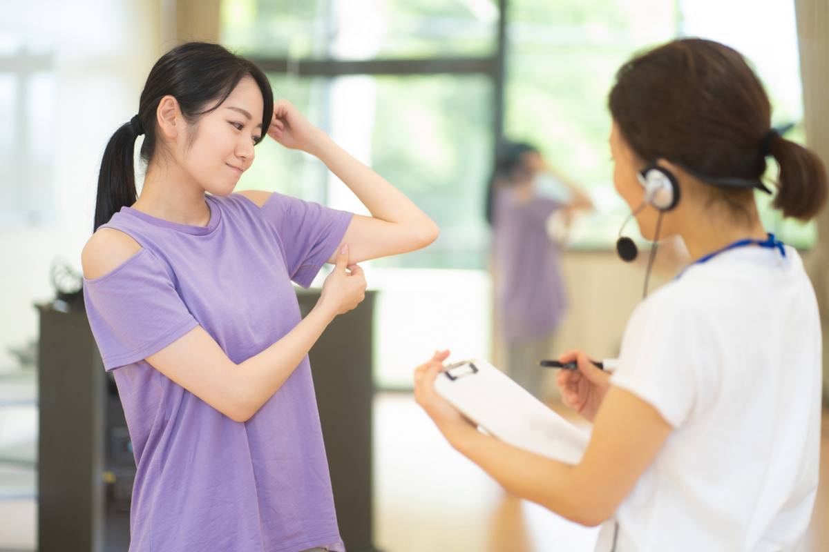 腕を胸の前で伸ばす女性の横で、スタッフがクリップボードに記録している様子
