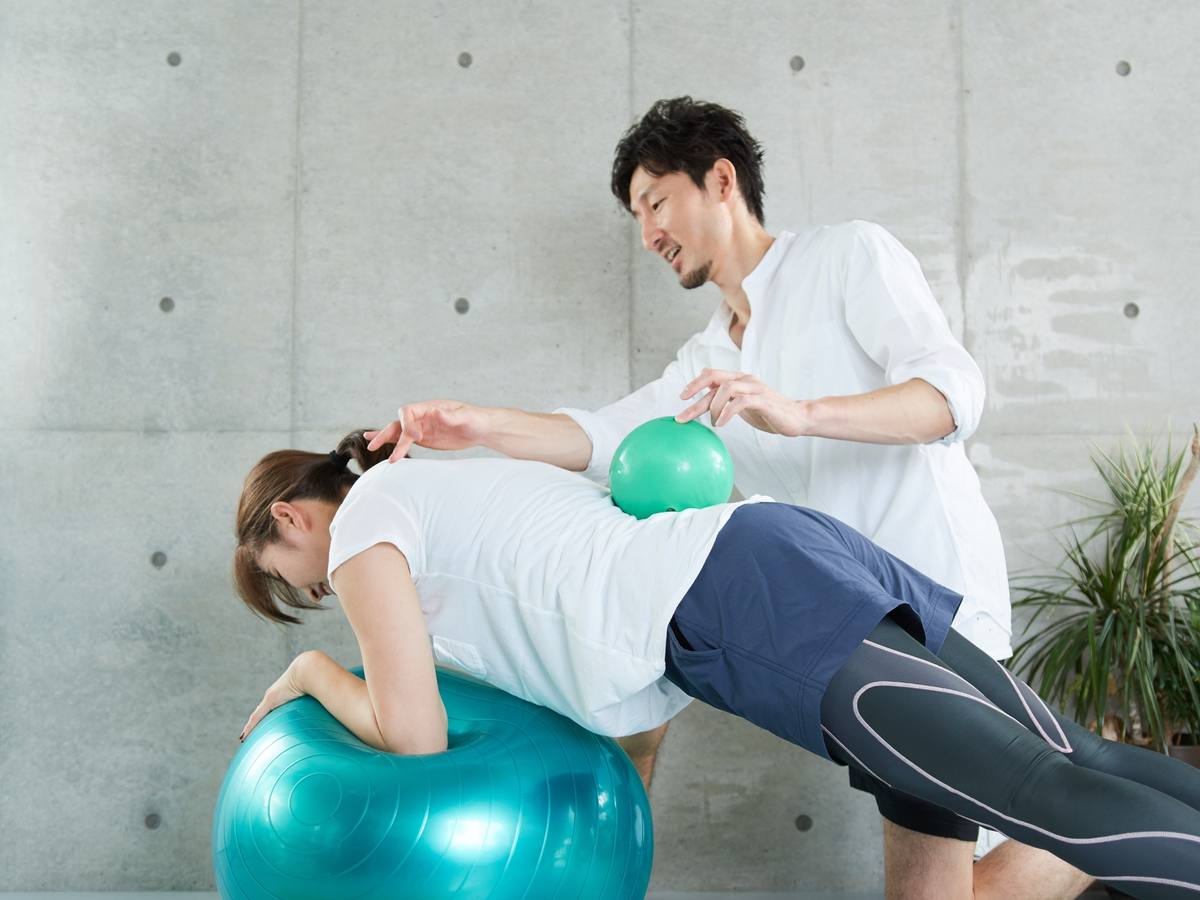 バランスボールにうつ伏せになった女性を、トレーナーが体を支えながら補助している様子