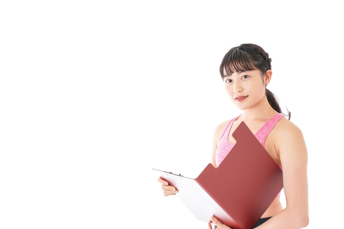 ピンクのウェアを着た女性トレーナーがクリップボードを手に持って立っている場面