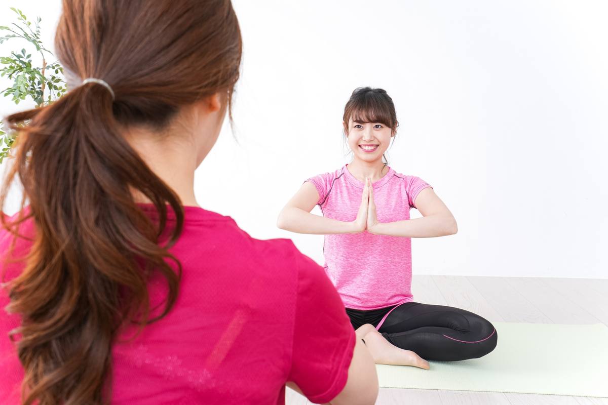白い部屋で、ピンクのウェアの女性の後ろに座り、胸の前で手を合わせる女性