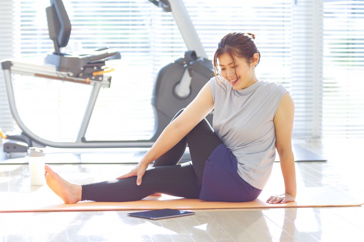 窓際のマットの上で、女性が脚を伸ばして上体をひねるストレッチをしている様子