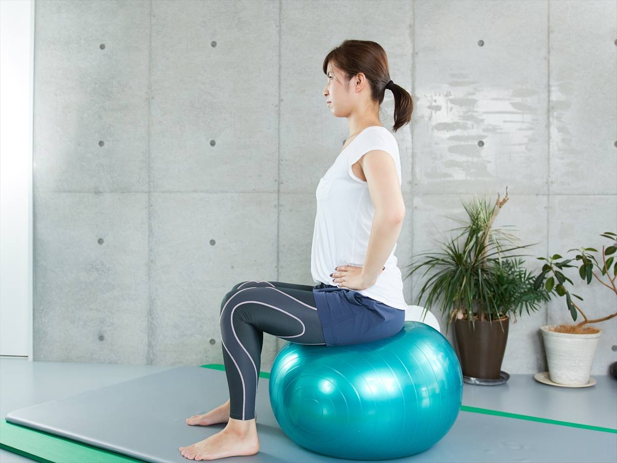 コンクリート壁の部屋でバランスボールに腰かけ、背筋を伸ばす女性を横から見た様子