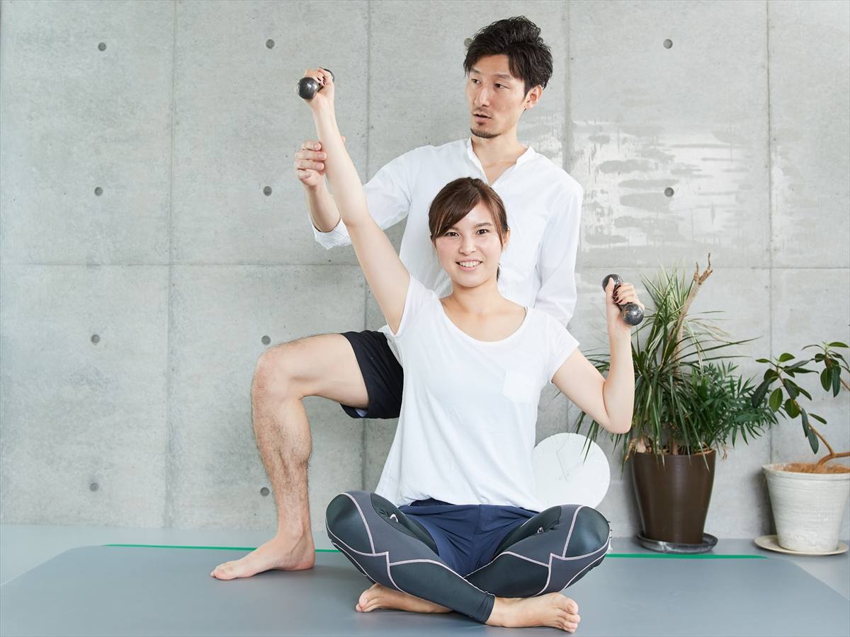 床に座ってダンベルを持つ女性の腕に、男性トレーナーが手を添えている様子