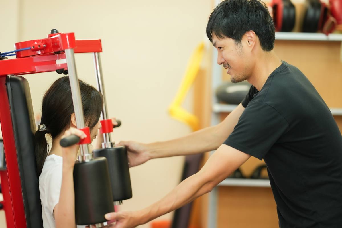 男性トレーナーが赤いマシンのパッドを調整し、女性の利用者が座って待っている場面
