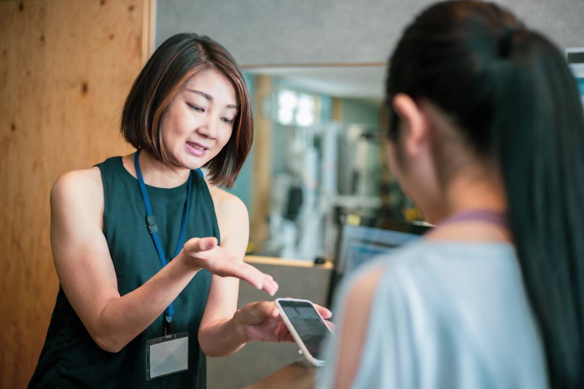 名札を付けた女性トレーナーがタブレットを手に、利用者へ身振りを交えて説明している場面
