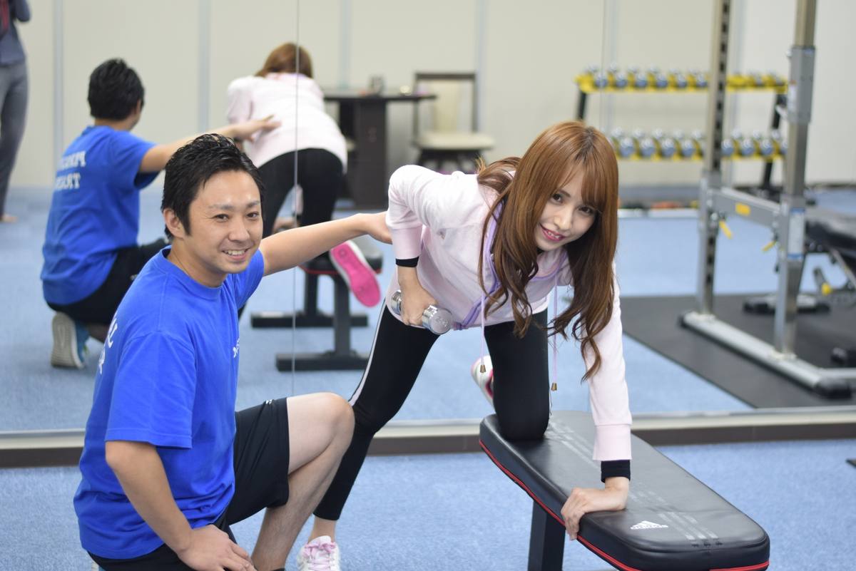 ステップ台を使う女性の動きを男性トレーナーが横で支えている場面