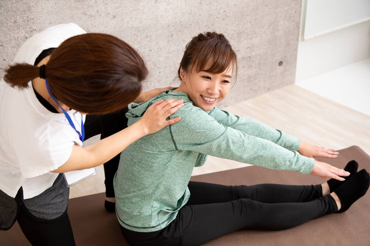床に座って両腕を前へ伸ばす女性の肩に、女性トレーナーが手を添えて支えている場面