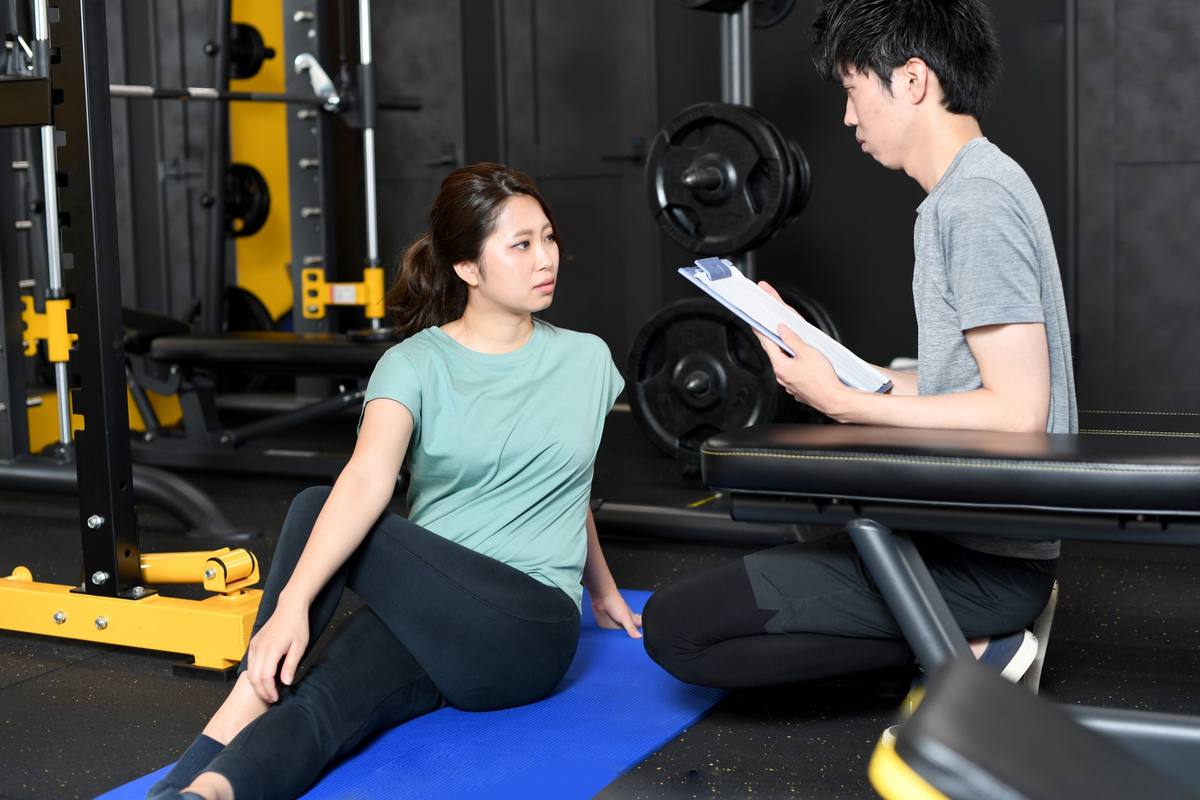 マットに座る女性利用者に、男性トレーナーがクリップボードを見せながら説明している様子