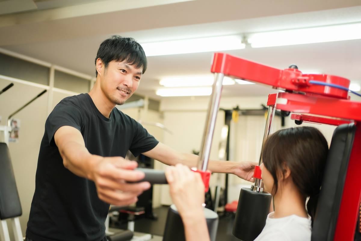 マシンの前でトレーナーが腕を伸ばし、座っている女性に動作を示している場面